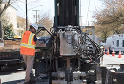 Pictured: A driller in a hi-viz vest and hard hat stands at the controls of a sonic drilling rig on a city street.